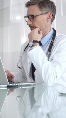 Kendine güvenen beyaz önlüklü erkek doktor laptopta çalışıyor, tıbbi verileri analiz ediyor ve modern hastane ortamında hasta bakımını planlıyor. İlaç ve sağlık hizmetleri