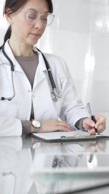 Beyaz önlüklü ve gözlüklü bir kadın doktor. Masada oturuyor, panoya yazıyor, hasta bilgilerini ve kayıtlarını yönetiyor. İlaç ve sağlık hizmetleri