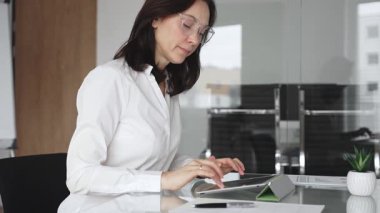 Bir telefon görüşmesi sırasında birden fazla iş yürüten işletme yöneticisi, çağdaş bir ofis ortamında temiz bir cam masadaki tablet ve küçük bir fabrika kullanarak
