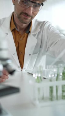 Laboratuvar önlüğü, beyaz eldiven ve koruyucu gözlük giyen bilim adamı, bir pipetten damlayan sıvıyı, içinde dikey yakın görüş olan yeşil bir bitki bulunan bir test tüpüne döküyor. Bilim ve tıp.