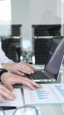 Bir dizüstü bilgisayarda iş birliği yapan ve mali tabloları inceleyen iş adamları, bir kişinin rapor üzerine notlar yazması, takım çalışması ve veri analizini göstermesi