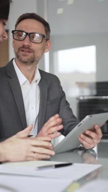 Kadın olarak masa başında işbirliği yapan iş adamları önemli bir sözleşme imzalarken, gözlüklü bir adam tablet üzerinde ayrıntılara dikkat çekiyor, takım çalışmasını ve karar verme mekanizmasını vurguluyor.