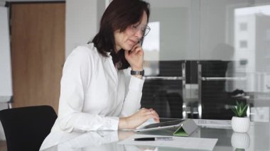 Beyaz bluzlu ve gözlüklü bir iş kadını modern bir ofis masasında oturuyor, ellerini çapraz yapıyor ve bir şirket toplantısı veya konferansı sırasında yoğun bir şekilde konsantre oluyor, yeni iş dünyasını derin bir şekilde düşünüyor.