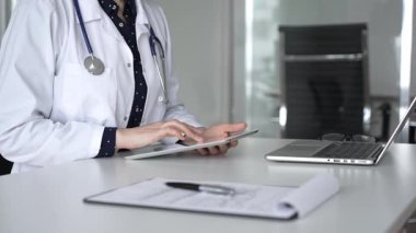 Modern dijital tablet kullanan beyaz önlüklü ve steteskoplu kadın doktor, klinik masasında hasta detayları üzerinde çalışıyor. Tıp, sağlık ve bilim konsepti