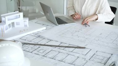 Mimari, inşaat planlaması için gerekli araçlarla çevrelenmiş, tasarım görevleri ve proje geliştirme için dizüstü bilgisayardan yararlanarak, fiziksel yapı modelinin yanında planları analiz eder.