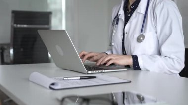 Beyaz önlüklü ve steteskoplu bir doktor dizüstü bilgisayarda daktilo kullanıyor, online sağlık kayıtlarını ve dijital tıbbi gelişmeleri yönetiyor. Tıp, sağlık ve bilim konsepti