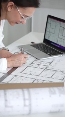 Kadın mimar, ayrıntılı bina planları üzerinde titizlikle çalışıyor. Masasına yayılmış. Mimari tasarımları parlak bir şekilde geliştirirken bir kalem ve cetvel kullanmak. Çağdaş ofis ortamı