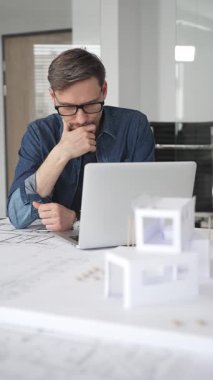 Profesyonel mimar, parlak bir ofis ortamında planlar ve dizüstü bilgisayarla çevriliyken bir masada tasarım çözümleri tasarlamaya odaklanır.