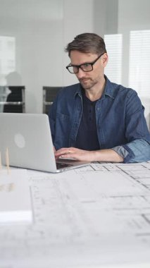 Profesyonel mimar, parlak bir ofis ortamında planlar ve dizüstü bilgisayarla çevriliyken bir masada tasarım çözümleri tasarlamaya odaklanır.