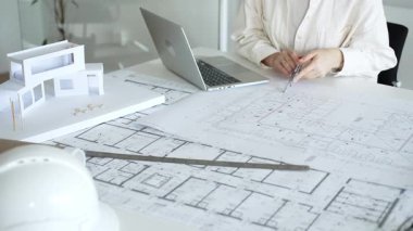 Mimari, inşaat planlaması için gerekli araçlarla çevrelenmiş, tasarım görevleri ve proje geliştirme için dizüstü bilgisayardan yararlanarak, fiziksel yapı modelinin yanında planları analiz eder.