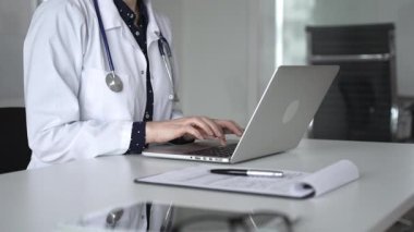 Beyaz önlüklü ve steteskoplu bir doktor dizüstü bilgisayarda daktilo kullanıyor, online sağlık kayıtlarını ve dijital tıbbi gelişmeleri yönetiyor. Tıp, sağlık ve bilim konsepti