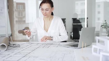 Kadın mimar, ayrıntılı bina planları üzerinde titizlikle çalışıyor. Masasına yayılmış. Mimari tasarımları parlak bir şekilde geliştirirken bir kalem ve cetvel kullanmak. Çağdaş ofis ortamı