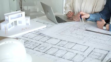 Birlikte çalışan mimarlar, bir dizüstü bilgisayar, mimari model ve güvenlik kaskıyla bir masaya yayılmış bina planlarını ölçüyor ve gözden geçiriyorlar, yeni inşaat gelişimine odaklanıyorlar.