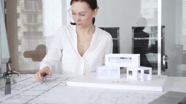 Masasında mimari planlar ve modeller üzerinde çalışan düşünceli bir kadın mimar elinde bir kalem tutuyor ve modern ofis ortamında proje detaylarını değerlendiriyor, kentsel gelişime odaklanıyor.