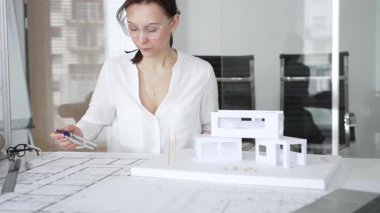 Masasında mimari planlar ve modeller üzerinde çalışan düşünceli bir kadın mimar elinde bir kalem tutuyor ve modern ofis ortamında proje detaylarını değerlendiriyor, kentsel gelişime odaklanıyor.