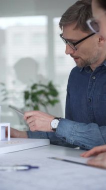 Mimari profesyoneller dizüstü bilgisayarlı bir masanın planlarını inceliyor, modern ofis ortamında tasarım kavramları ve proje geliştirme üzerine odaklanmış bir tartışmaya giriyorlar.