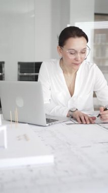 Kadın mimar, hassas ve profesyonel odaklı yenilikçi bir konut tasarımını ölçmek ve arıtmak için araçlar kullanarak, masa başında çağdaş yapı modeli ve planlarını inceler.