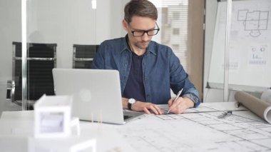 Profesyonel mimar modern bir masadaki detaylı yerleşim planlarını detaylı bir şekilde gözden geçirir. Laptop ve taslak araçları kullanarak kusursuz planlar yapar. Çağdaş bir ofiste teknolojiyle ev tasarımları yapılır.