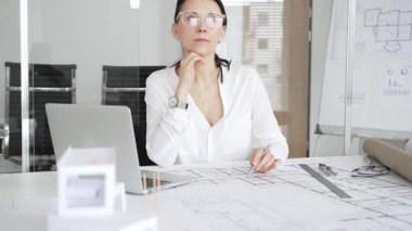 Masasında mimari planlar ve modeller üzerinde çalışan düşünceli bir kadın mimar elinde bir kalem tutuyor ve modern ofis ortamında proje detaylarını değerlendiriyor, kentsel gelişime odaklanıyor.