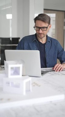 Erkek mimar masasına odaklanmış, dizüstü bilgisayardaki planları ve dijital planları gözden geçiriyor, modern ofis ortamında yenilikçi yerleşim ve kentsel tasarım çözümleri geliştiriyor.