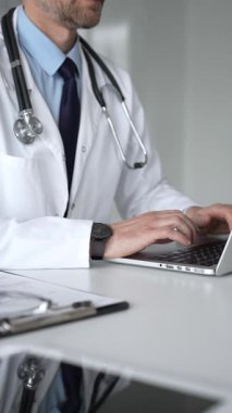 Beyaz önlüklü ve steteskoplu bir doktor gümüş bir dizüstü bilgisayarda yazı yazıyor, online tıbbi konsültasyon sağlıyor ve hasta kayıtlarını yönetiyor. Tıp, sağlık ve bilim konsepti