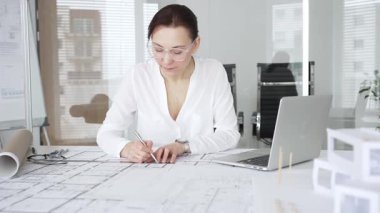 Kadın mimar, ayrıntılı bina planları üzerinde titizlikle çalışıyor. Masasına yayılmış. Mimari tasarımları parlak bir şekilde geliştirirken bir kalem ve cetvel kullanmak. Çağdaş ofis ortamı