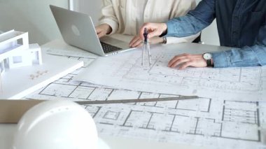 Birlikte çalışan mimarlar, bir dizüstü bilgisayar, mimari model ve güvenlik kaskıyla bir masaya yayılmış bina planlarını ölçüyor ve gözden geçiriyorlar, yeni inşaat gelişimine odaklanıyorlar.