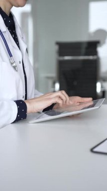 Modern dijital tablet kullanan beyaz önlüklü ve steteskoplu kadın doktor, klinik masasında hasta detayları üzerinde çalışıyor. Tıp, sağlık ve bilim konsepti