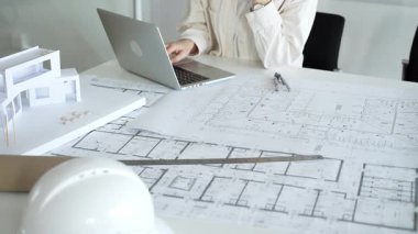 Mimari, inşaat planlaması için gerekli araçlarla çevrelenmiş, tasarım görevleri ve proje geliştirme için dizüstü bilgisayardan yararlanarak, fiziksel yapı modelinin yanında planları analiz eder.