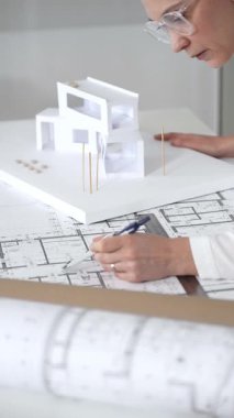 Kadın mimar, ayrıntılı bina planları üzerinde titizlikle çalışıyor. Masasına yayılmış. Mimari tasarımları parlak bir şekilde geliştirirken bir kalem ve cetvel kullanmak. Çağdaş ofis ortamı