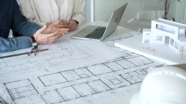 Birlikte çalışan mimarlar, bir dizüstü bilgisayar, mimari model ve güvenlik kaskıyla bir masaya yayılmış bina planlarını ölçüyor ve gözden geçiriyorlar, yeni inşaat gelişimine odaklanıyorlar.