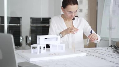 Masasında mimari planlar ve modeller üzerinde çalışan düşünceli bir kadın mimar elinde bir kalem tutuyor ve modern ofis ortamında proje detaylarını değerlendiriyor, kentsel gelişime odaklanıyor.