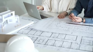 Birlikte çalışan mimarlar, bir dizüstü bilgisayar, mimari model ve güvenlik kaskıyla bir masaya yayılmış bina planlarını ölçüyor ve gözden geçiriyorlar, yeni inşaat gelişimine odaklanıyorlar.