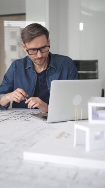 Profesyonel mimar, parlak bir ofis ortamında planlar ve dizüstü bilgisayarla çevriliyken bir masada tasarım çözümleri tasarlamaya odaklanır.