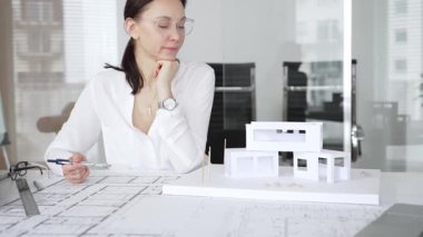 Masasında mimari planlar ve modeller üzerinde çalışan düşünceli bir kadın mimar elinde bir kalem tutuyor ve modern ofis ortamında proje detaylarını değerlendiriyor, kentsel gelişime odaklanıyor.