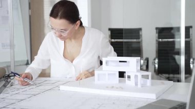 Masasında mimari planlar ve modeller üzerinde çalışan düşünceli bir kadın mimar elinde bir kalem tutuyor ve modern ofis ortamında proje detaylarını değerlendiriyor, kentsel gelişime odaklanıyor.
