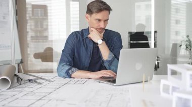 Profesyonel mimar modern bir masadaki detaylı yerleşim planlarını detaylı bir şekilde gözden geçirir. Laptop ve taslak araçları kullanarak kusursuz planlar yapar. Çağdaş bir ofiste teknolojiyle ev tasarımları yapılır.