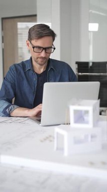 Profesyonel mimar, parlak bir ofis ortamında planlar ve dizüstü bilgisayarla çevriliyken bir masada tasarım çözümleri tasarlamaya odaklanır.