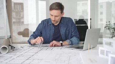 Profesyonel mimar modern bir masadaki detaylı yerleşim planlarını detaylı bir şekilde gözden geçirir. Laptop ve taslak araçları kullanarak kusursuz planlar yapar. Çağdaş bir ofiste teknolojiyle ev tasarımları yapılır.