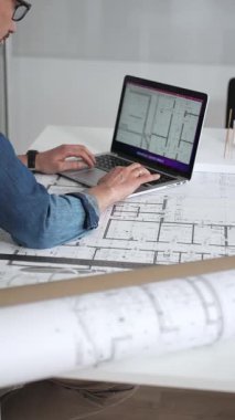 Mimar, yeni projeler için ayrıntılı planlama, inşaat ve tasarım mühendisliğini sembolize eden, kat planlarını gösteren bir cetvel ve dizüstü bilgisayar kullanarak beyaz bir masanın üzerine bina planları çiziyor.