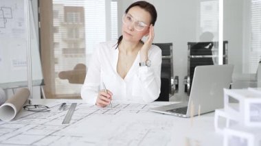 Masasında mimari planlar ve modeller üzerinde çalışan düşünceli bir kadın mimar elinde bir kalem tutuyor ve modern ofis ortamında proje detaylarını değerlendiriyor, kentsel gelişime odaklanıyor.