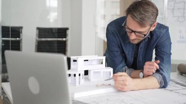 Düzensiz bir masada ayrıntılı planlar hazırlayan mimar planlar, planlar, dizüstü bilgisayarlar ve kağıt model evleri, konut ve ticari tasarım ve geliştirme projelerine odaklanmış.