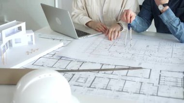 Birlikte çalışan mimarlar, bir dizüstü bilgisayar, mimari model ve güvenlik kaskıyla bir masaya yayılmış bina planlarını ölçüyor ve gözden geçiriyorlar, yeni inşaat gelişimine odaklanıyorlar.