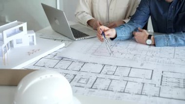 Birlikte çalışan mimarlar, bir dizüstü bilgisayar, mimari model ve güvenlik kaskıyla bir masaya yayılmış bina planlarını ölçüyor ve gözden geçiriyorlar, yeni inşaat gelişimine odaklanıyorlar.