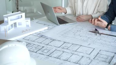 Birlikte çalışan mimarlar, bir dizüstü bilgisayar, mimari model ve güvenlik kaskıyla bir masaya yayılmış bina planlarını ölçüyor ve gözden geçiriyorlar, yeni inşaat gelişimine odaklanıyorlar.