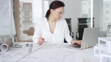 Kadın mimar, ayrıntılı bina planları üzerinde titizlikle çalışıyor. Masasına yayılmış. Mimari tasarımları parlak bir şekilde geliştirirken bir kalem ve cetvel kullanmak. Çağdaş ofis ortamı