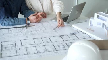 Birlikte çalışan mimarlar, bir dizüstü bilgisayar, mimari model ve güvenlik kaskıyla bir masaya yayılmış bina planlarını ölçüyor ve gözden geçiriyorlar, yeni inşaat gelişimine odaklanıyorlar.