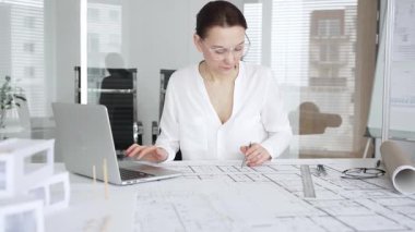 Kadın mimar, ayrıntılı bina planları üzerinde titizlikle çalışıyor. Masasına yayılmış. Mimari tasarımları parlak bir şekilde geliştirirken bir kalem ve cetvel kullanmak. Çağdaş ofis ortamı