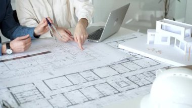 Birlikte çalışan mimarlar, bir dizüstü bilgisayar, mimari model ve güvenlik kaskıyla bir masaya yayılmış bina planlarını ölçüyor ve gözden geçiriyorlar, yeni inşaat gelişimine odaklanıyorlar.
