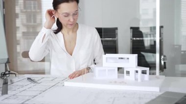 Masasında mimari planlar ve modeller üzerinde çalışan düşünceli bir kadın mimar elinde bir kalem tutuyor ve modern ofis ortamında proje detaylarını değerlendiriyor, kentsel gelişime odaklanıyor.